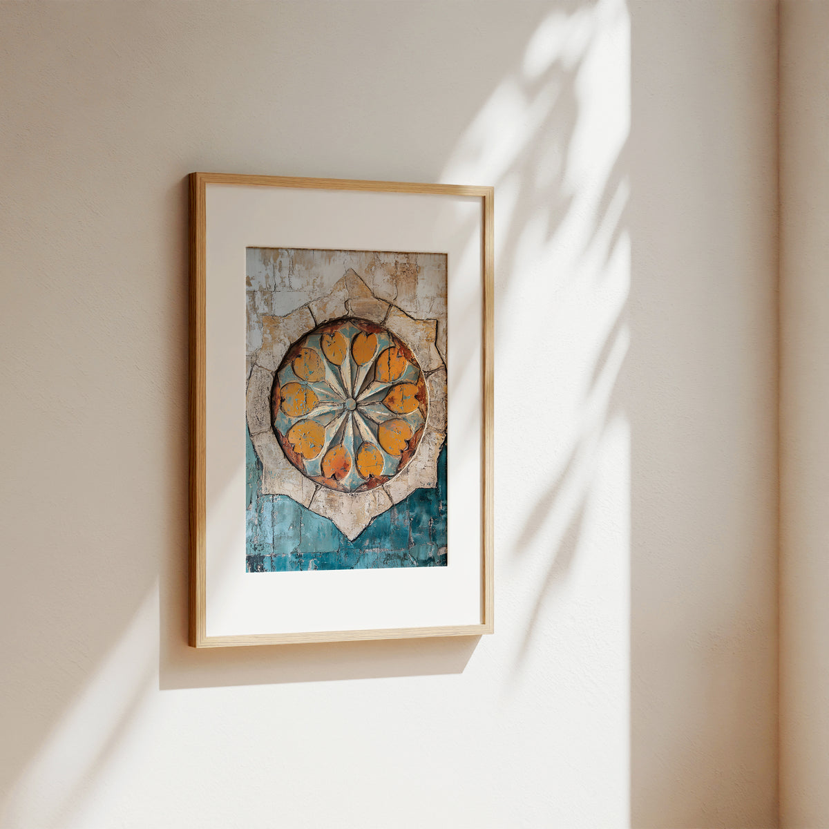 A framed art print featuring a circular mandala design in orange, yellow, and blue tones, mounted on a beige wall with soft sunlight and leaf shadows.