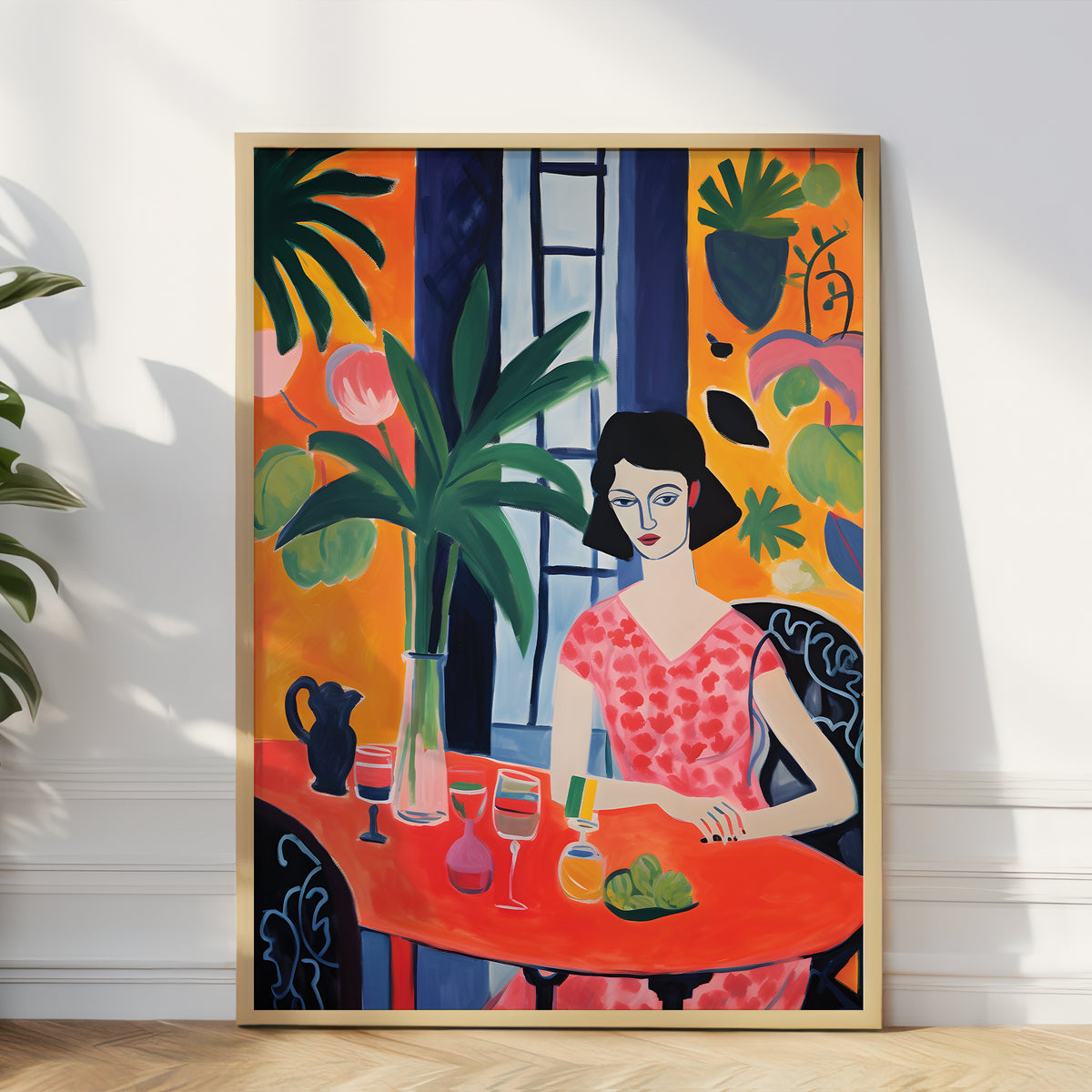 A framed colorful art print leaning against a white wall on a light wood floor, showing a stylized woman seated at an orange table with plants and bold geometric background elements.