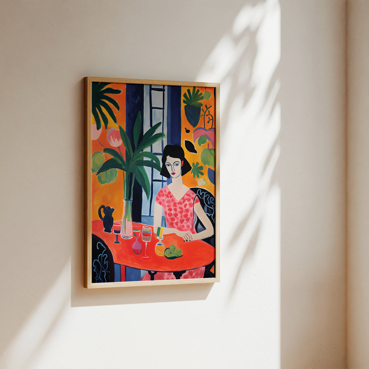 A framed colorful art print depicting a stylized woman seated at a table with tropical plants, hung on a beige wall with soft sunlight and leaf shadows cast across it.