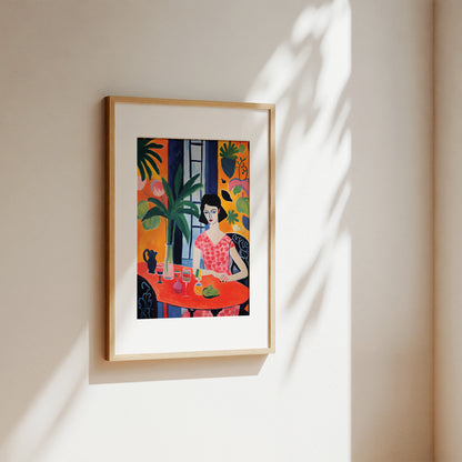 A framed art print featuring a colorful, stylized illustration of a woman sitting at a table with plants, hanging on a beige wall with soft sunlight and leaf shadows.