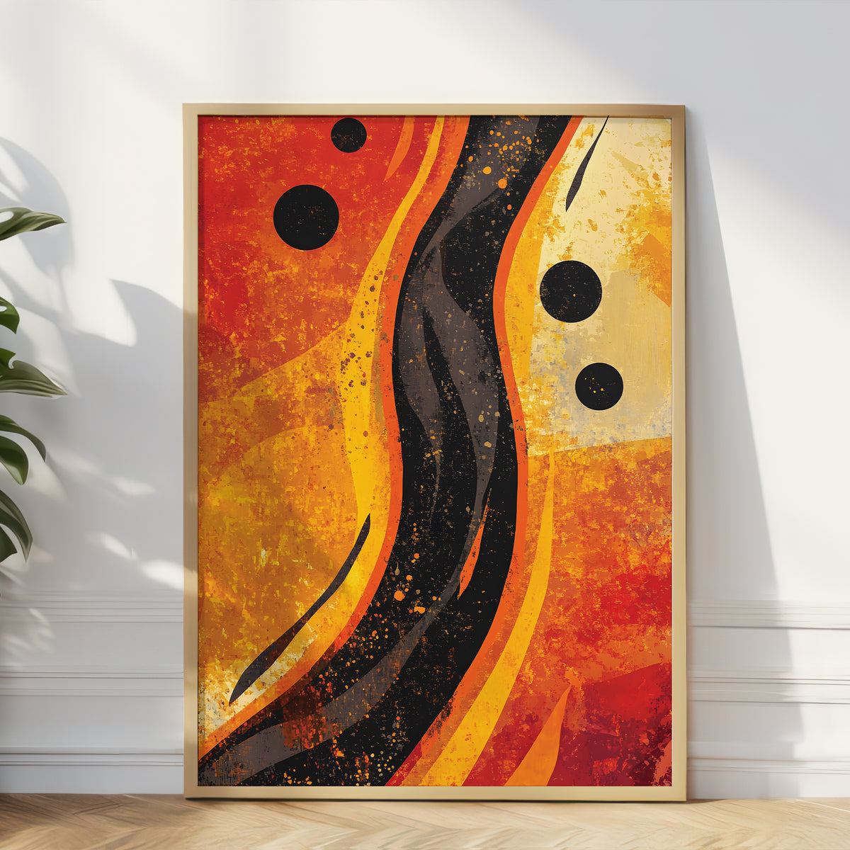 A framed abstract wall art print leaning against a white wall in a room. The artwork features bold, flowing waves and splatters in orange, yellow, black, and red, enclosed in a light wood frame.