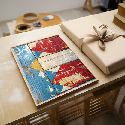 A framed rustic wooden art print with a distressed paint pattern in red, blue, beige, and yellow, displayed on a light wood table next to a wrapped gift box with a beige ribbon.