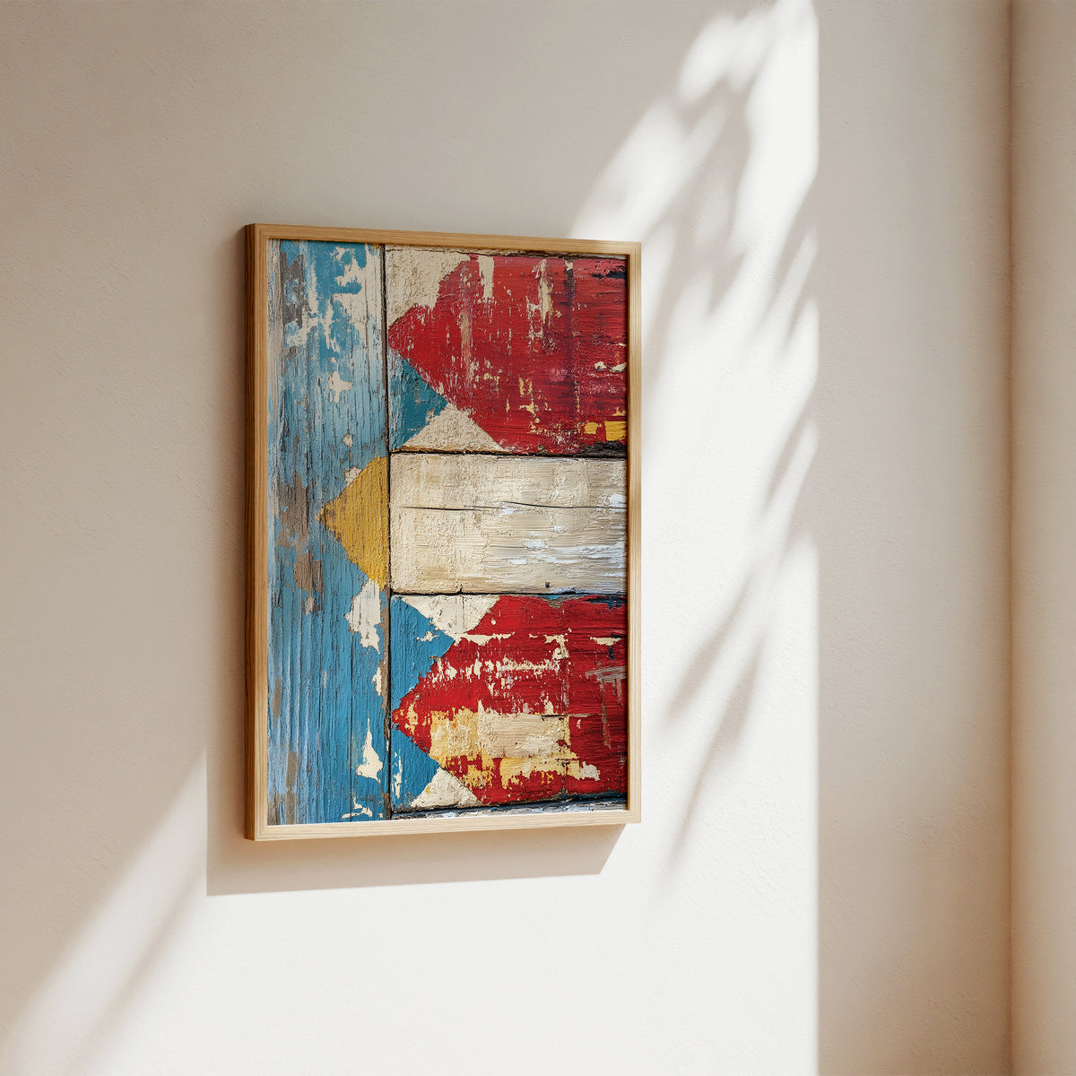 A framed abstract art print featuring a distressed, weathered design with red, blue, cream, and yellow panels, hanging on a warm beige wall with soft sunlight and leaf shadows.