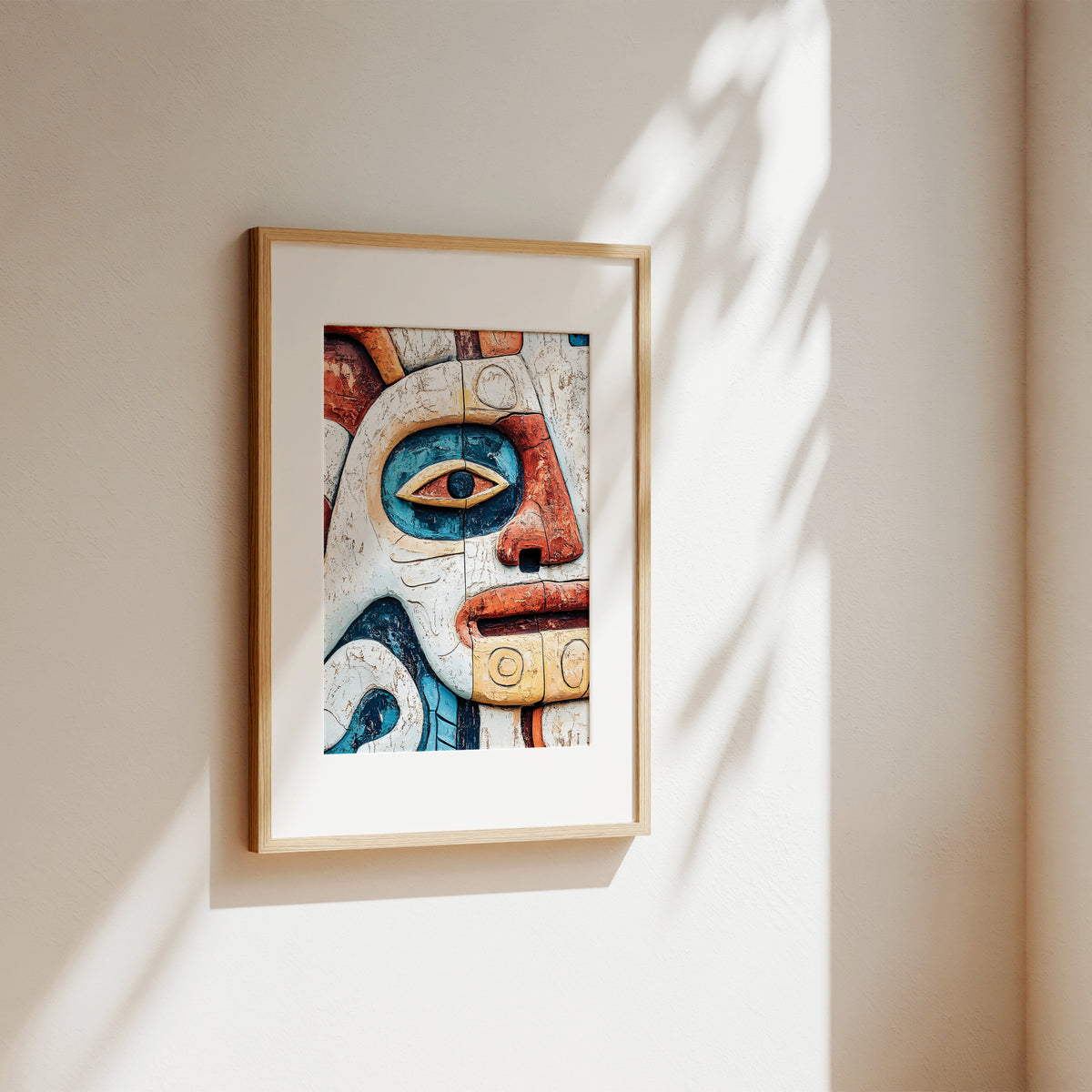A framed art print featuring a colorful, stylized indigenous face design, hung on a warm beige wall with soft, dappled sunlight casting leaf shadows.