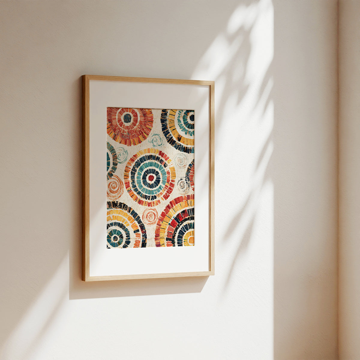 A framed art print featuring colorful circular mosaic patterns, hung on a warm beige wall with soft sunlight and leaf shadows casting across it.