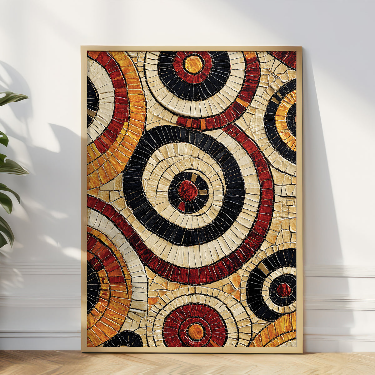 Framed abstract wall art featuring concentric circular mosaic-style patterns in cream, black, red, orange, and brown, leaning against a white wall on a wood floor with a plant partially visible at left.