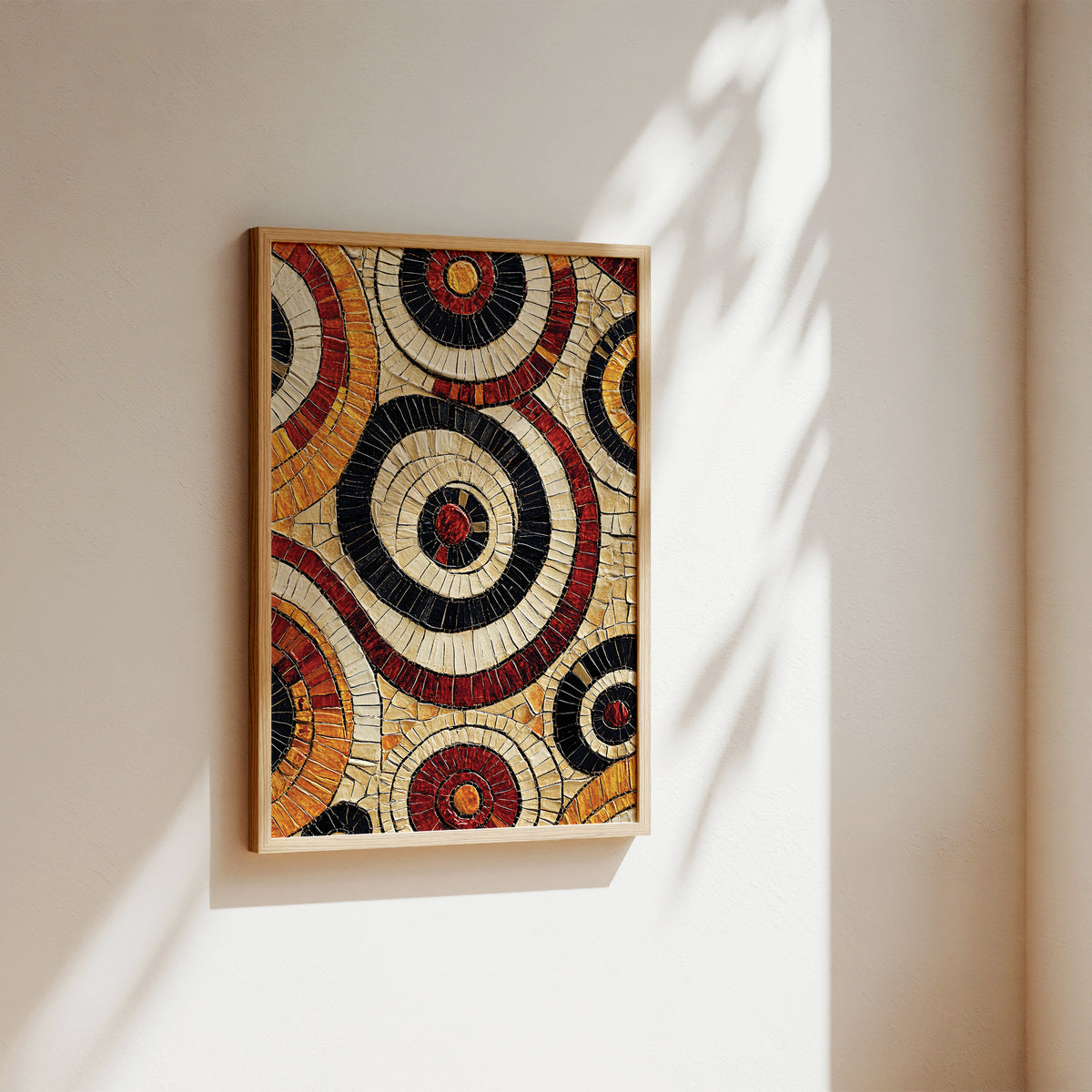 A framed circular mosaic wall art piece with concentric rings in cream, black, red, and orange, hanging on a warm beige wall with soft sunlight and leaf shadows.