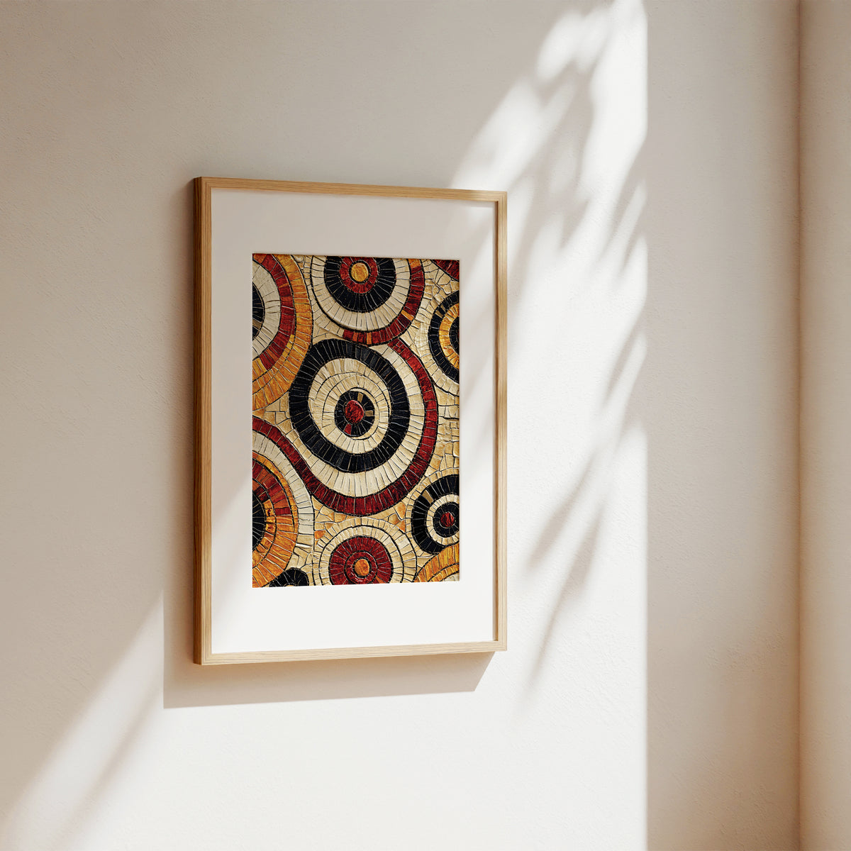 A framed art print featuring a mosaic design of concentric circles in black, red, gold, and cream, displayed on a warm beige wall with soft sunlight and leaf shadows.