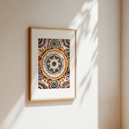 A framed art print featuring a detailed, colorful circular mandala design, hanging on a warm beige wall with soft sunlight and leaf shadows cast across it.