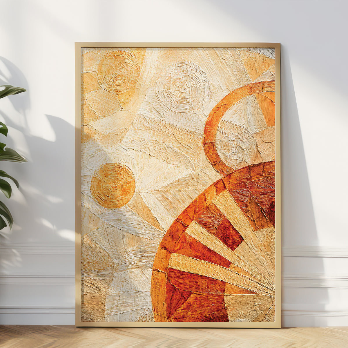A framed abstract art print leaning against a white wall on a light wood floor, with a plant leaf visible at the left edge. The artwork features warm cream, tan, orange, and rust tones in a textured, layered composition inside a light wood frame.