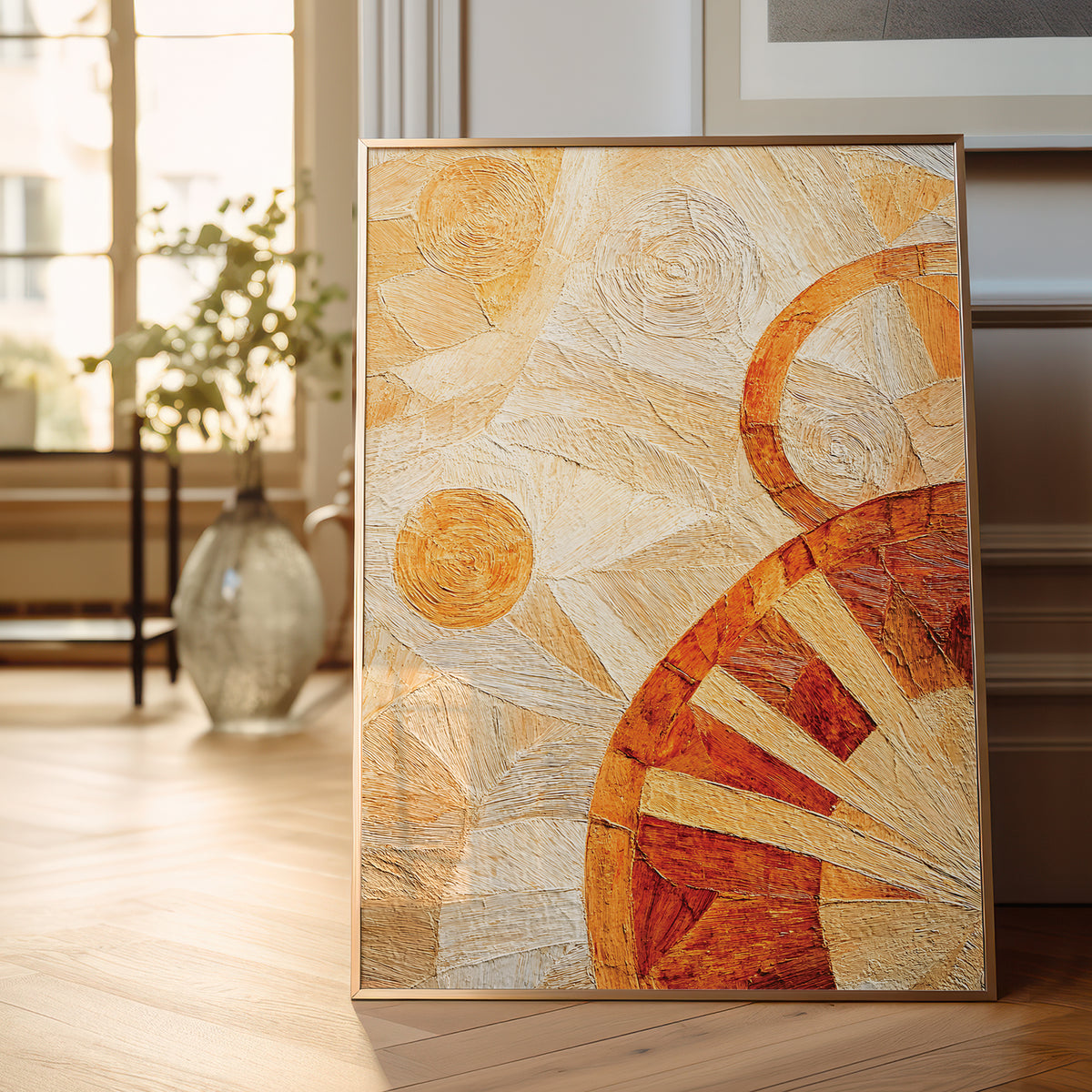 A framed abstract canvas print leaning against a wall on a light wood floor in a bright, modern room. The artwork features textured brushstrokes in warm tones of orange, terracotta, cream, and beige, with geometric and sun-like circular motifs.