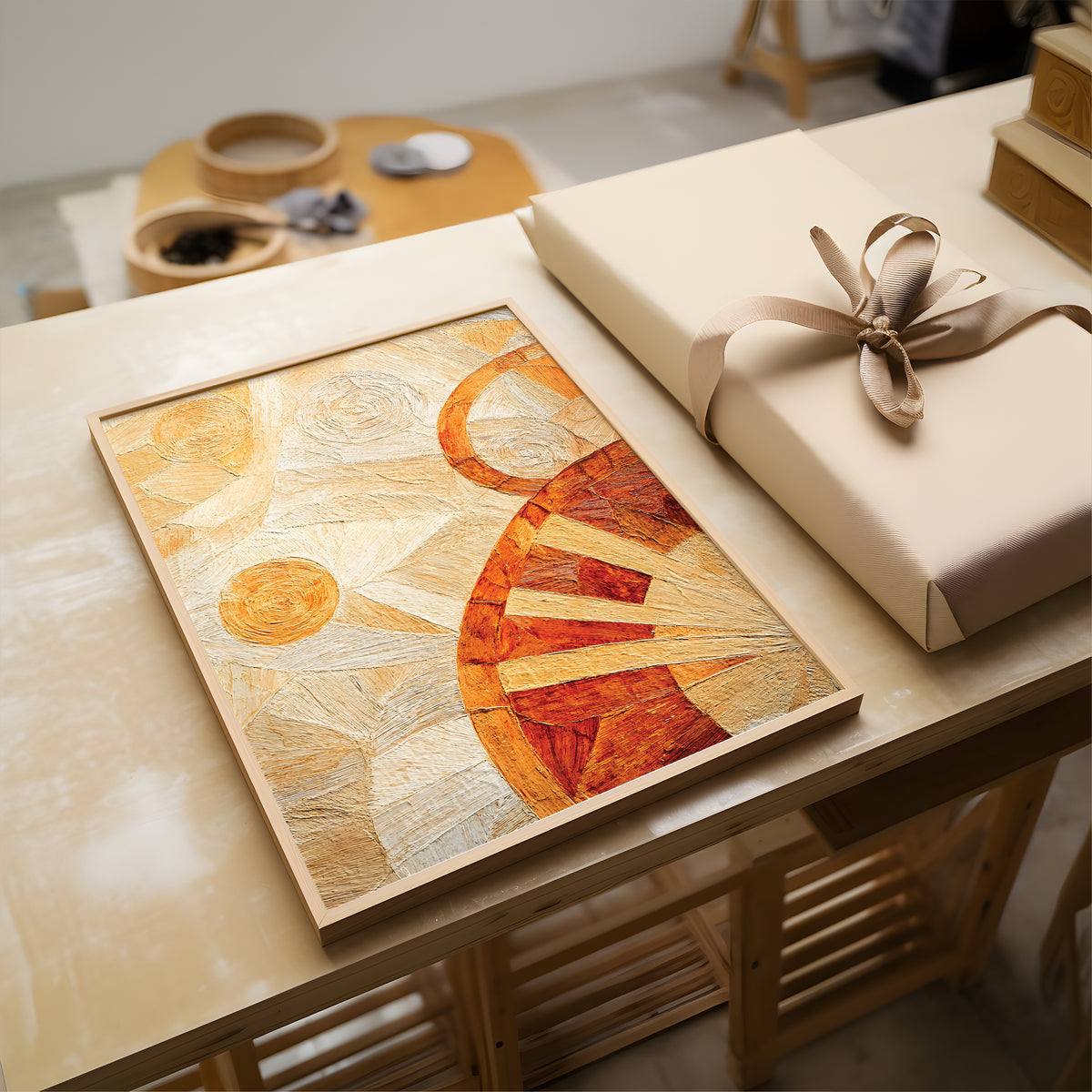 A framed abstract art print with warm orange, red, and beige geometric patterns, displayed on a wooden table next to a wrapped gift box with a beige ribbon.