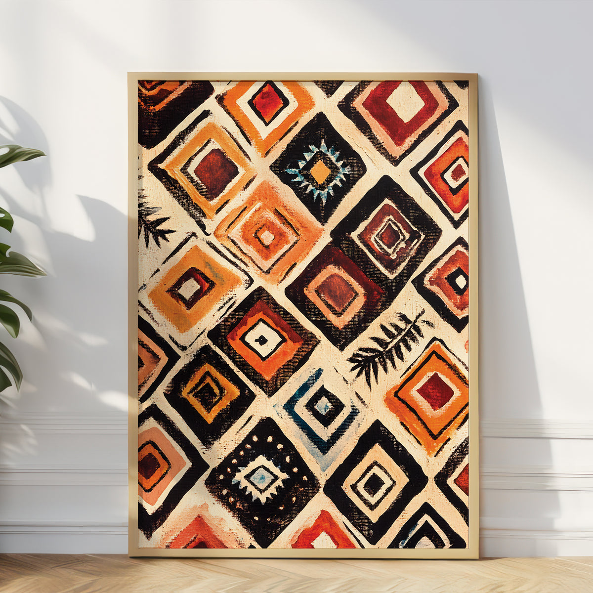 A framed abstract geometric wall art print with a bold diamond pattern in orange, red, black, and cream, leaning against a white wall on a light wood floor next to a green plant.