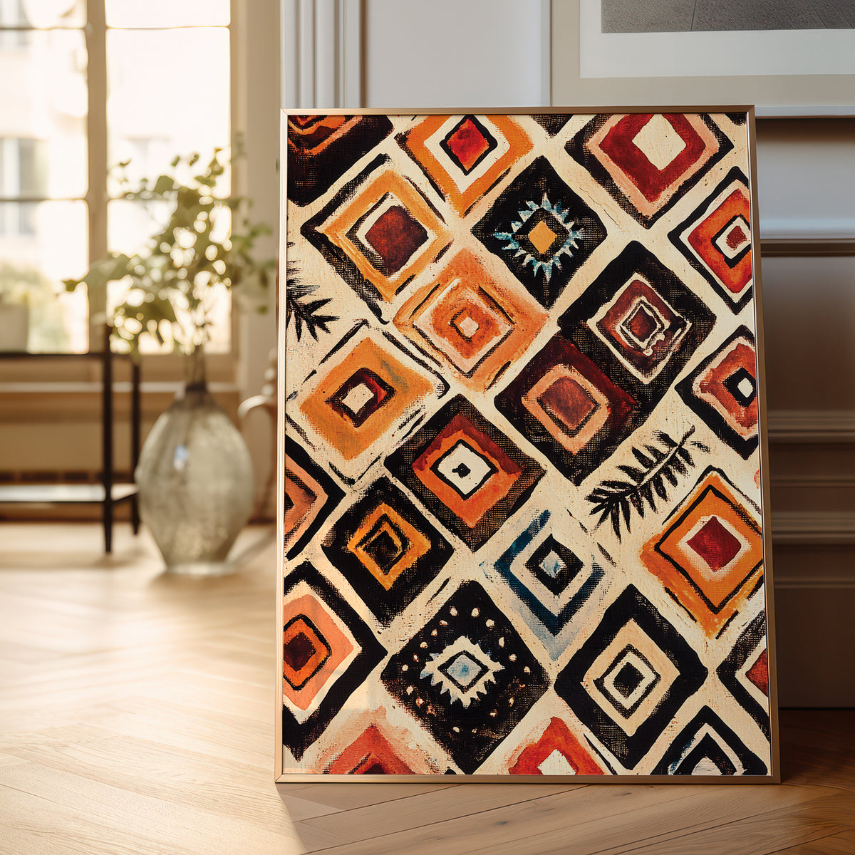 A framed geometric canvas print with a bold diamond pattern in orange, red, black, and cream, leaning against a wall on a light wood floor in a bright room.