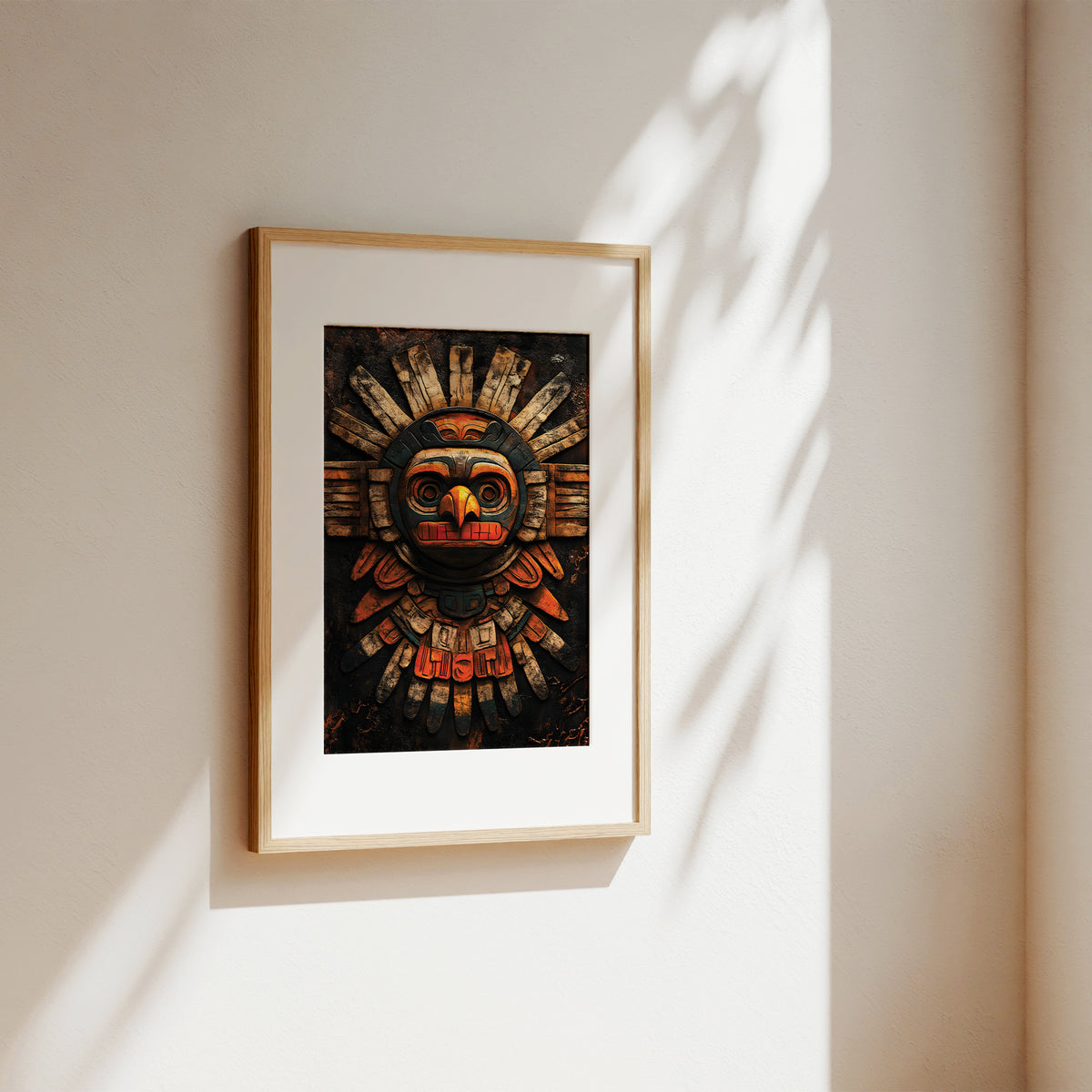 A framed art print featuring a stylized, colorful indigenous-style mask on a dark background, displayed on a warm beige wall with soft sunlight and leaf shadows.