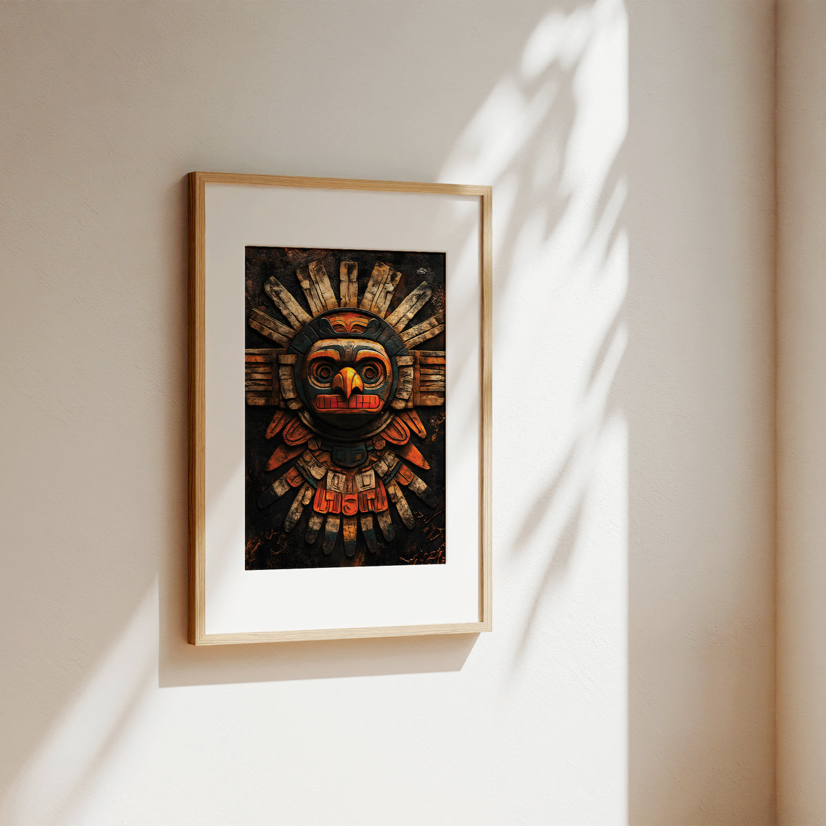 A framed art print featuring a stylized, colorful indigenous-style mask on a dark background, displayed on a warm beige wall with soft sunlight and leaf shadows.