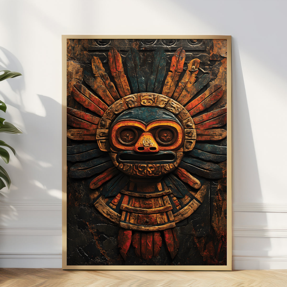 A framed vertical art print featuring an Aztec-style sun mask with dark teal, rust red, and gold-brown tones, leaning against a white wall on a light wood floor with a plant at the side.