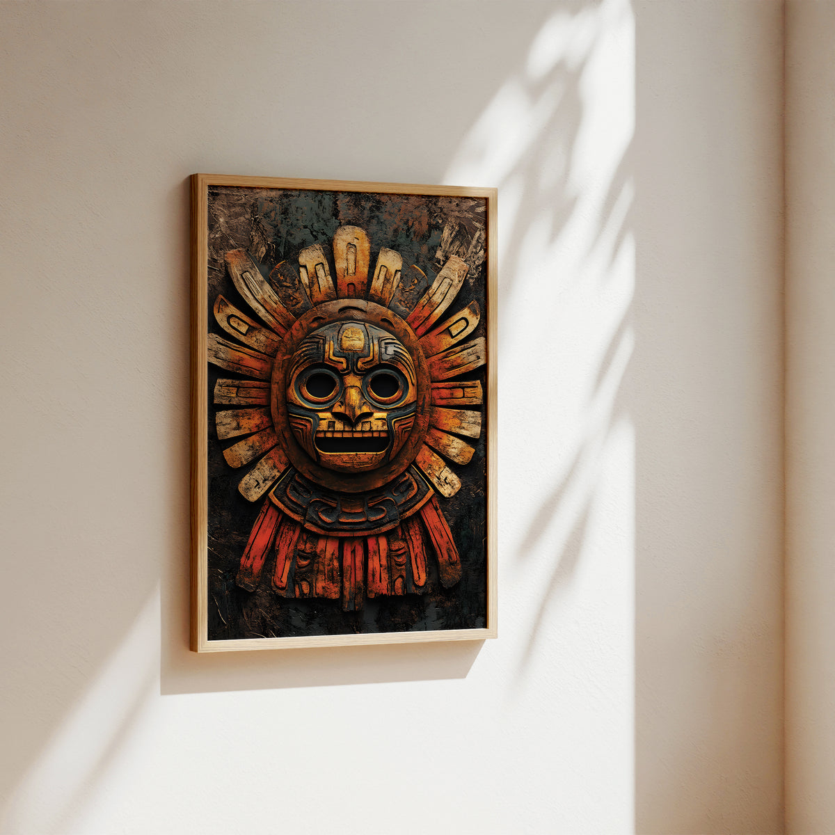 A framed art print featuring an Aztec-style sun mask with gold, red, and black details, hanging on a warm beige wall with soft sunlight and leaf shadows.