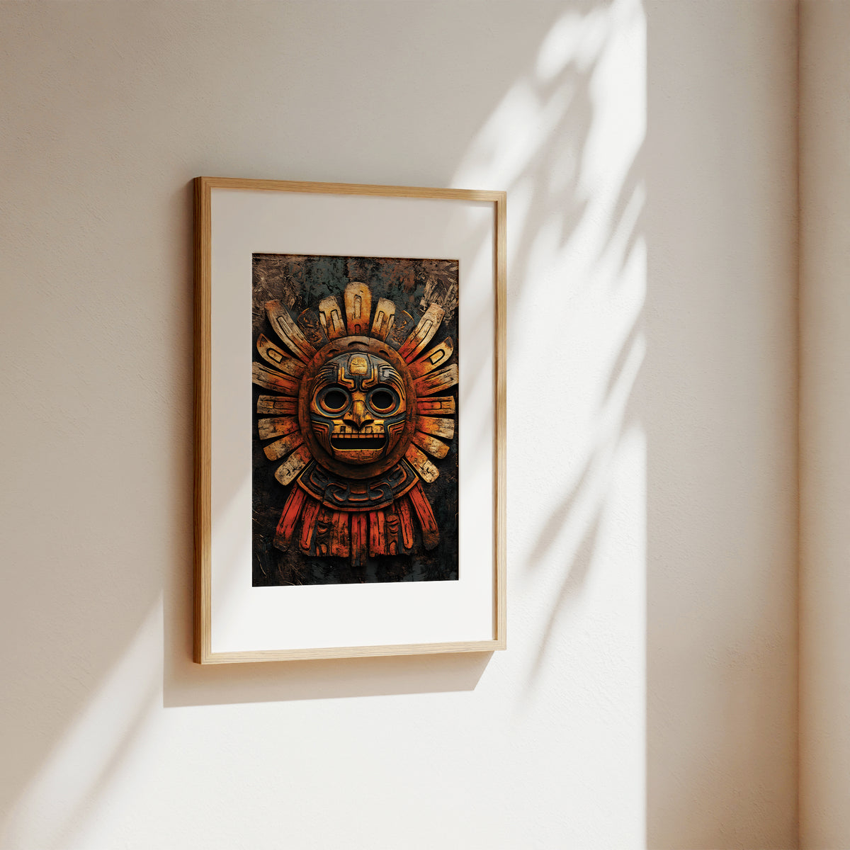 A framed art print featuring a detailed Aztec sun mask, displayed on a warm beige wall with soft, natural leaf shadows cast across it.