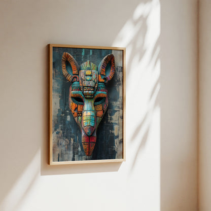 A framed wall art print featuring a colorful stylized ram or goat head mask, hung on a beige wall with soft sunlight and leaf shadows cast across it.