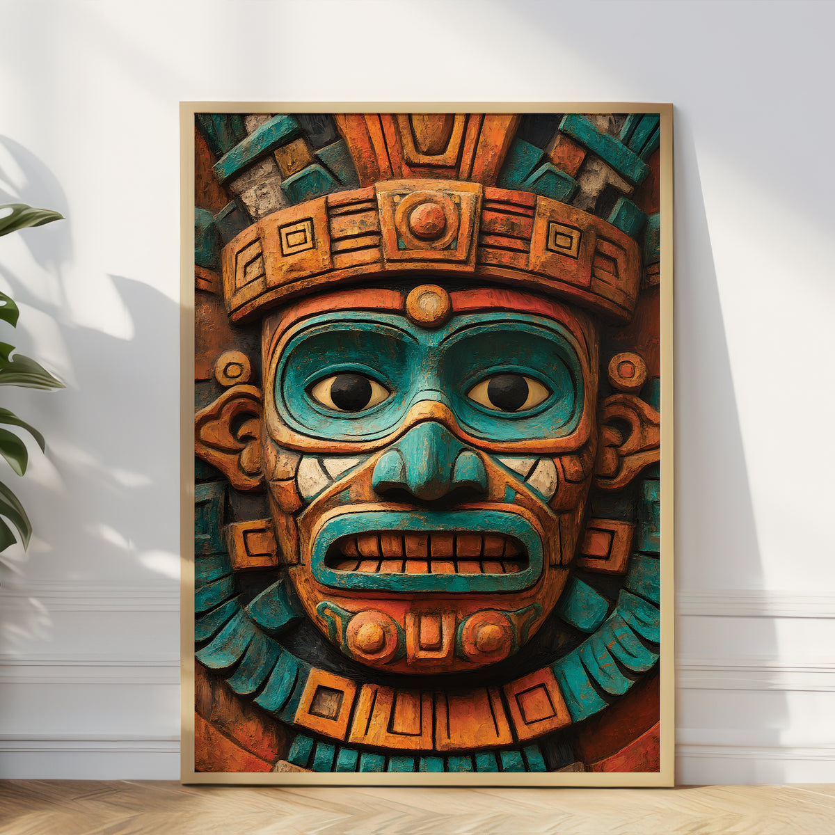 A framed art print featuring a detailed Aztec-style carved face mask in vibrant turquoise and terracotta orange, leaning against a white wall on a light wood floor next to a green plant.