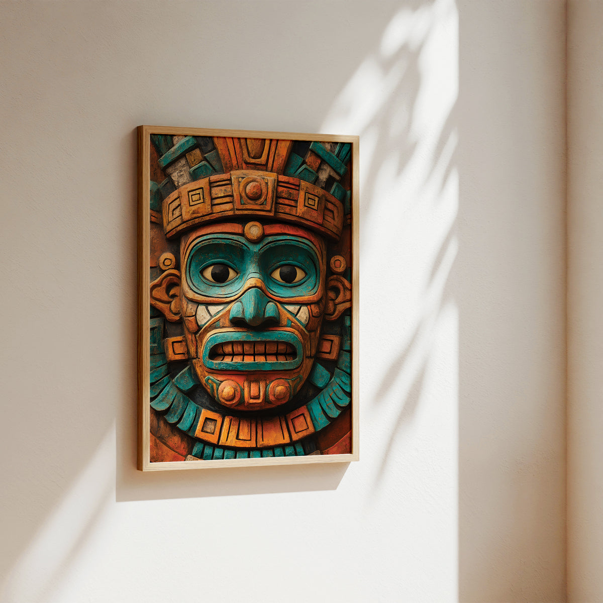 A framed art print featuring a detailed Aztec-style mask in vibrant turquoise, orange, and brown tones, hanging on a warm beige wall with soft sunlight and leaf shadows.