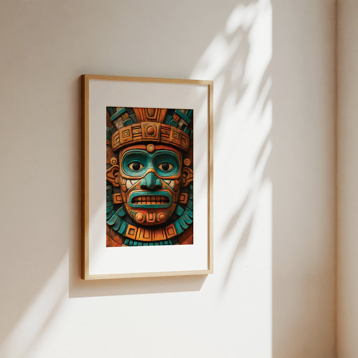 A framed art print featuring a detailed Aztec-style face mask in teal, gold, and brown tones, displayed on a warm beige wall with soft sunlight and leaf shadows.