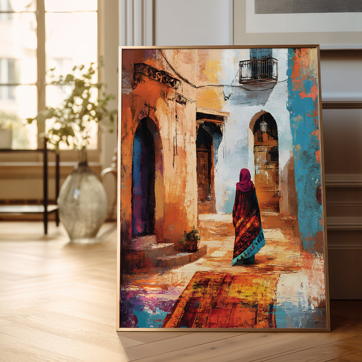 A framed painting leaning against a wall on a wooden floor in a bright room. The artwork depicts a narrow sunlit alley with arched doorways, warm ochre and beige walls, blue accents, and a woman in a magenta headscarf walking away.