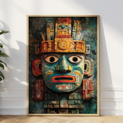 A framed vertical art print featuring a stylized Aztec-inspired face mask in teal, gold, red, and brown tones, leaning against a white wall on a light wood floor with a plant partially visible at the left edge.