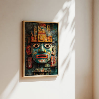 A framed art print featuring a detailed Aztec-style calendar mask in vibrant teal, gold, and red tones, hanging on a warm beige wall with soft diagonal sunlight and leaf shadows.
