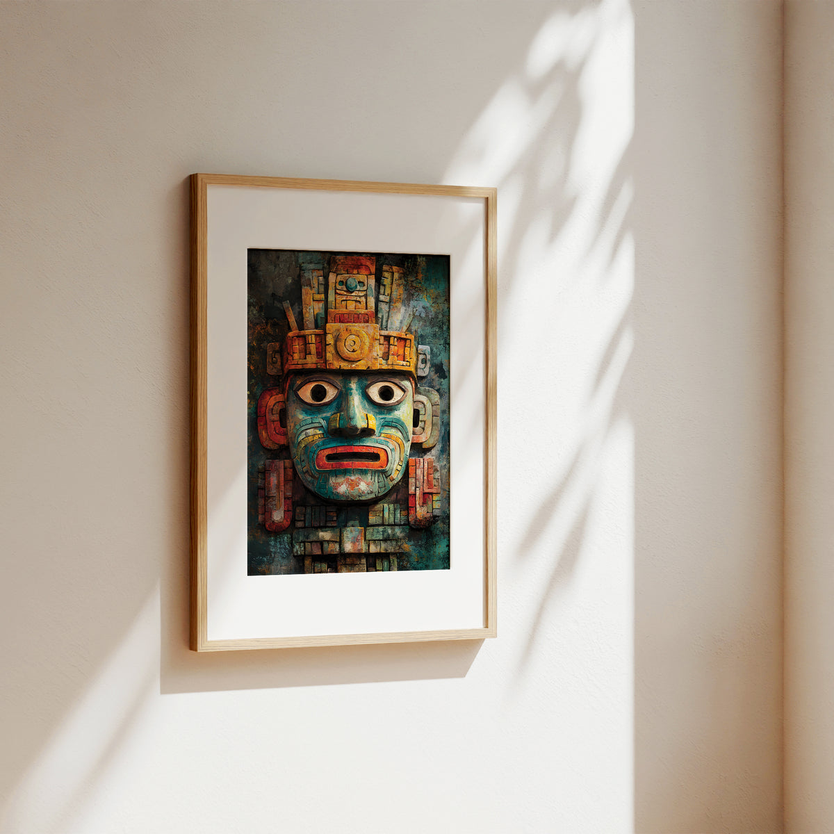 A framed art print featuring a detailed Aztec-style face mask with turquoise, red, and gold tones, displayed on a warm beige wall with soft leaf shadows.