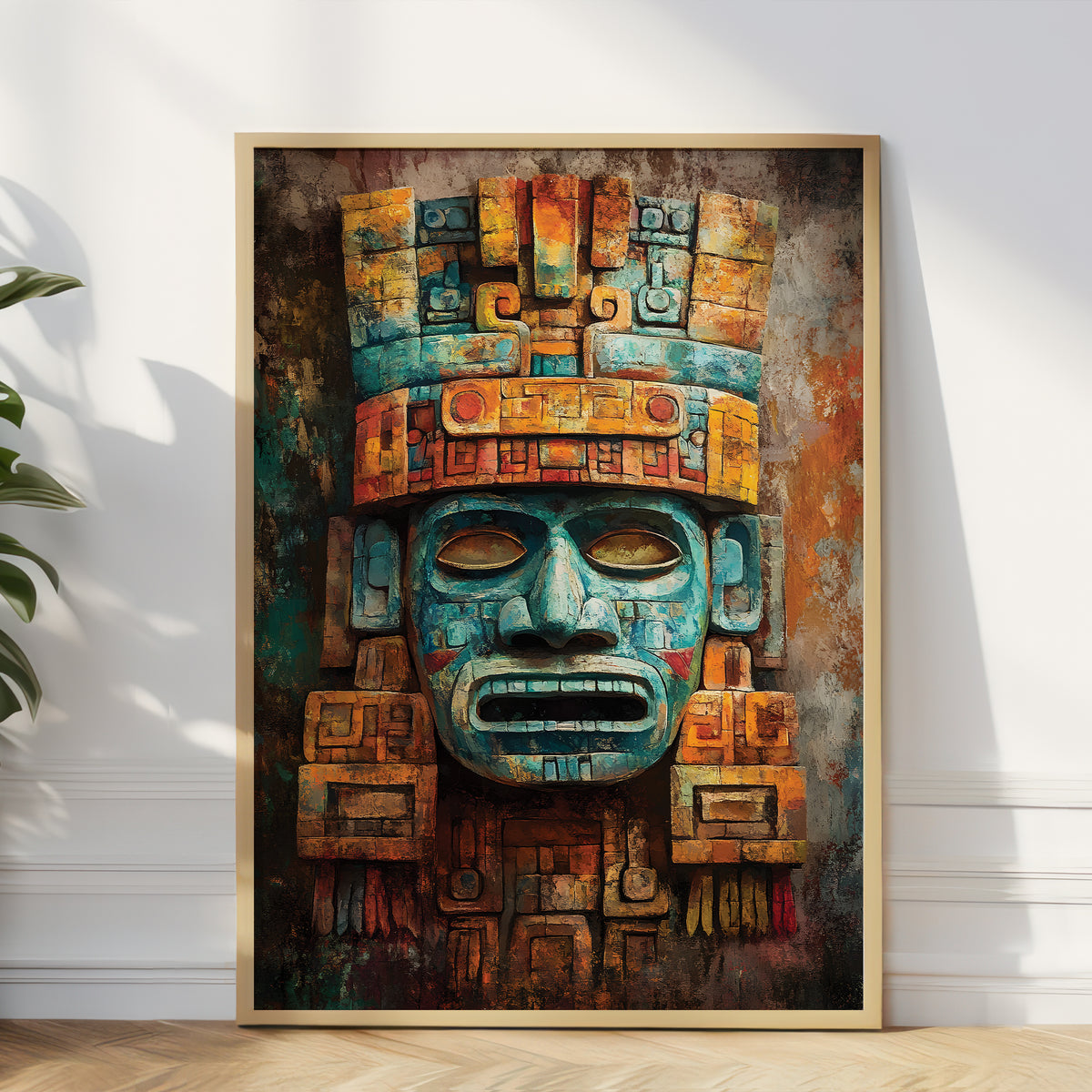 A framed vertical art print featuring a stylized Aztec-style face mask in teal, orange, brown, and gold tones, leaning against a white wall on a light wood floor with a plant partially visible at the left edge.