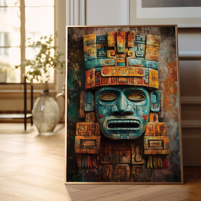 A framed vertical art print featuring a stylized Aztec-style face mask in teal, gold, and brown tones, leaning against a wall on a light wood floor in a softly lit interior room.