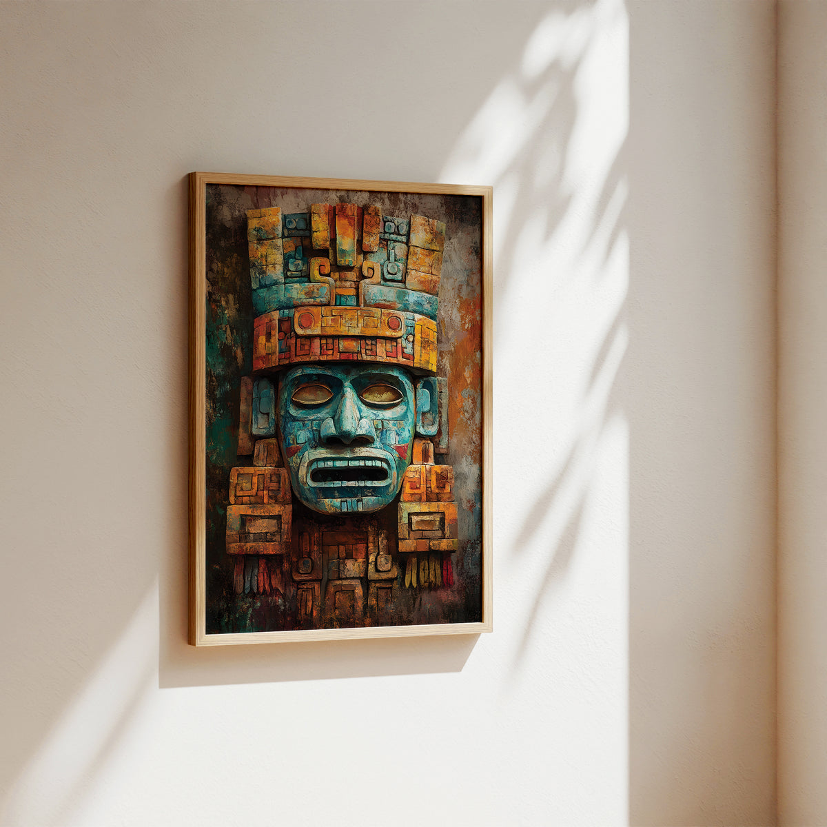 A framed wall art print featuring a stylized Aztec-style face mask in teal, orange, and brown tones, hanging on a beige wall with soft sunlight and leaf shadows.
