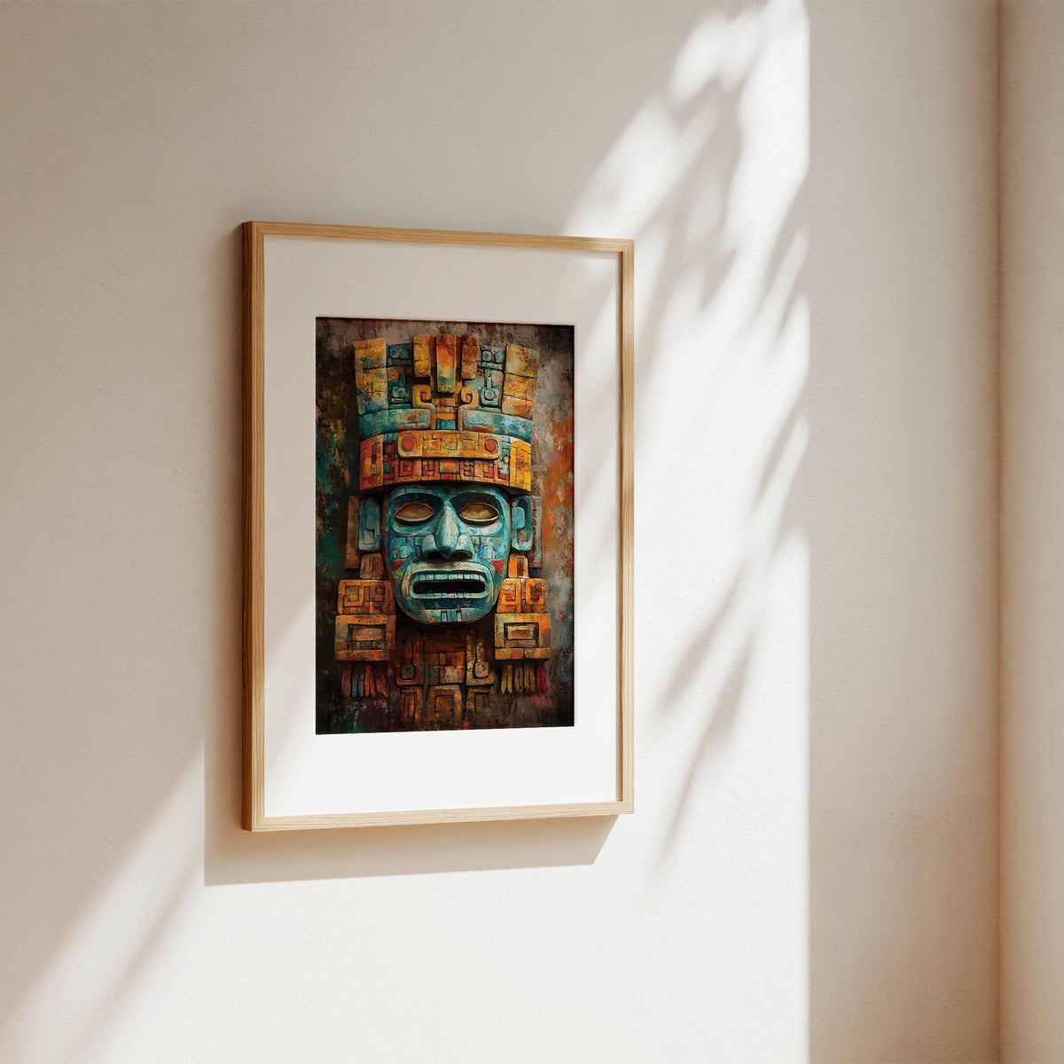 A framed art print featuring a detailed Aztec-style face mask in teal, gold, and brown tones, displayed on a warm beige wall with soft leaf shadows cast across it.