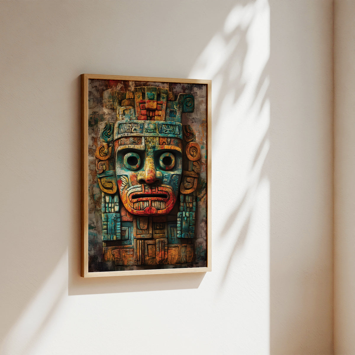 A framed art print featuring a detailed Aztec-style calendar mask in vibrant teal, red, and gold tones, hanging on a warm beige wall with soft sunlight and leaf shadows.