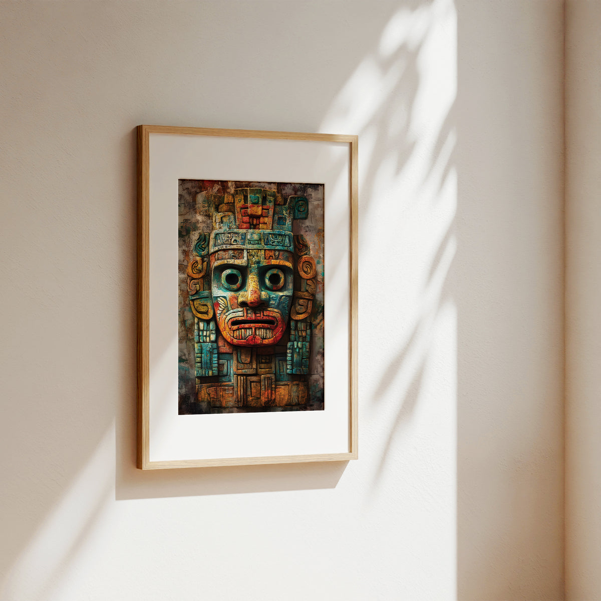 A framed art print featuring a detailed Aztec-style face mask, displayed on a warm beige wall with soft, natural leaf shadows cast across it.