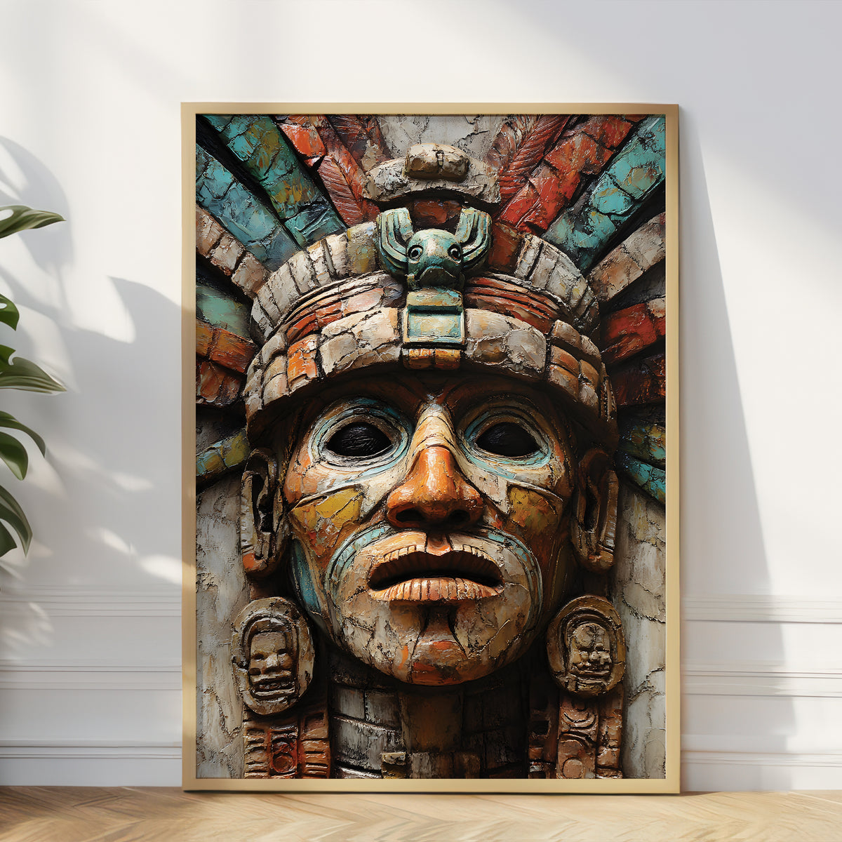 A large framed art print leaning against a white wall on a light wood floor, showing a detailed Aztec-style face mask with a feathered headdress in earthy multicolor tones. A plant and cast window shadows are visible on the left and right of the scene.