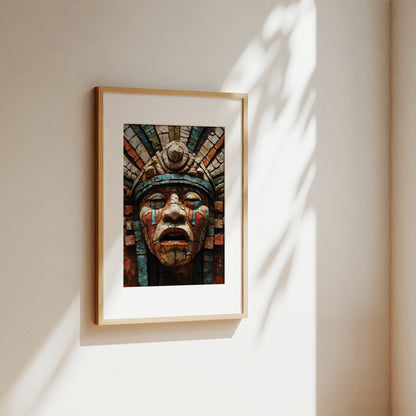 A framed art print featuring an Aztec-style face sculpture in earthy red, blue, and brown tones, hung on a warm beige wall with soft sunlight and leaf shadows.