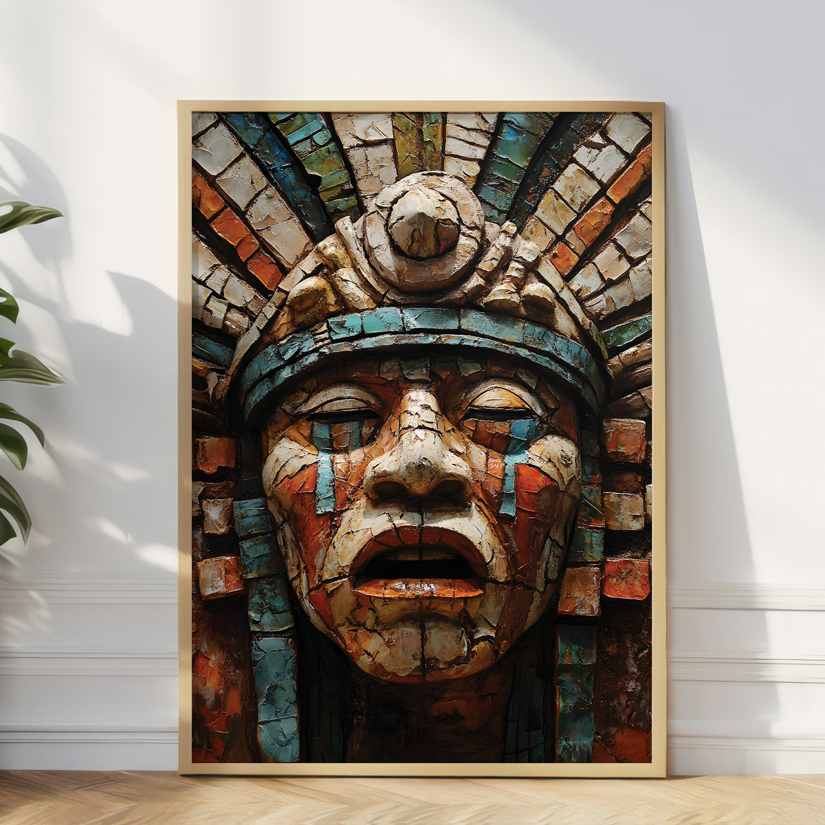 A large framed wall art print leaning against a white wall on a wood floor, showing a close-up of an Aztec-style face with a colorful mosaic headdress. The print has a light wood frame and is styled in a modern room setting with a plant on the left.