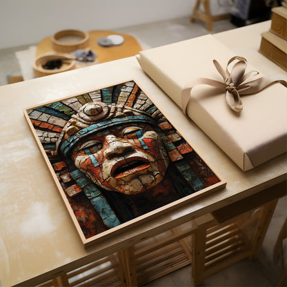 A framed art print featuring an Aztec-style mosaic face mask in earthy red, blue, and beige tones, displayed on a light wood table next to a wrapped gift box with a beige ribbon.