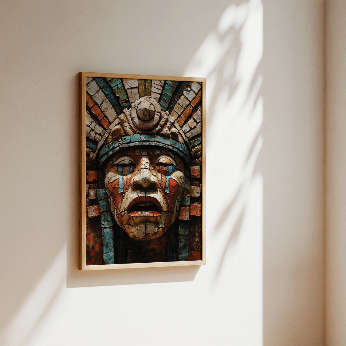 A framed art print featuring a detailed Aztec-style face sculpture with a colorful mosaic background, hanging on a warm beige wall with soft sunlight and leaf shadows.