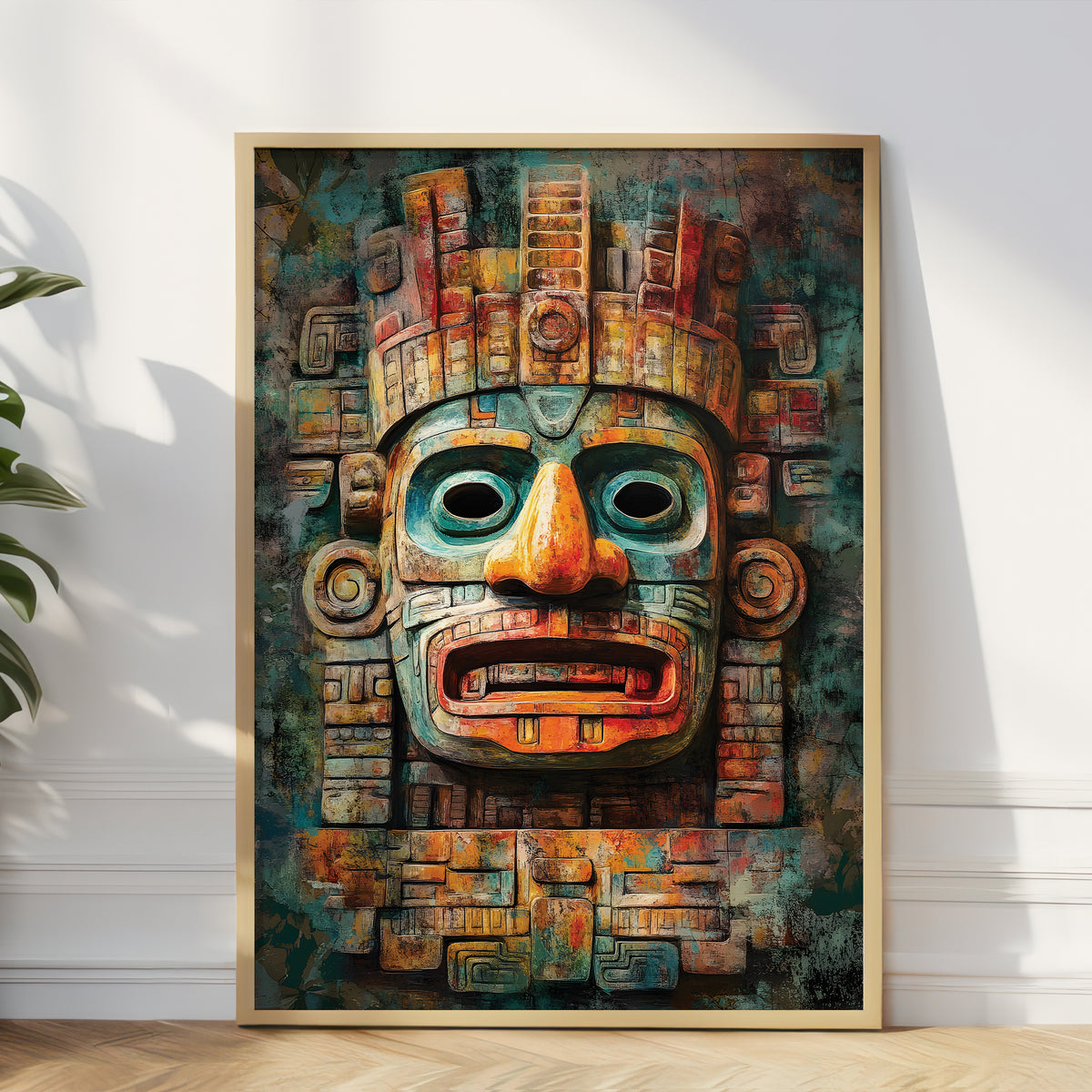 A framed vertical art print featuring a stylized Aztec-style mask face in teal, rust orange, and brown tones, leaning against a white wall on a light wood floor with a plant partially visible at the left edge.