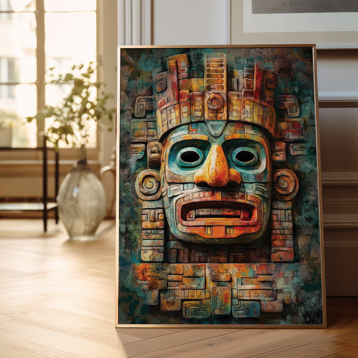 A framed art print featuring a stylized Aztec-style mask with teal, orange, and brown tones, leaning against a wall on a light wood floor in a softly lit room.