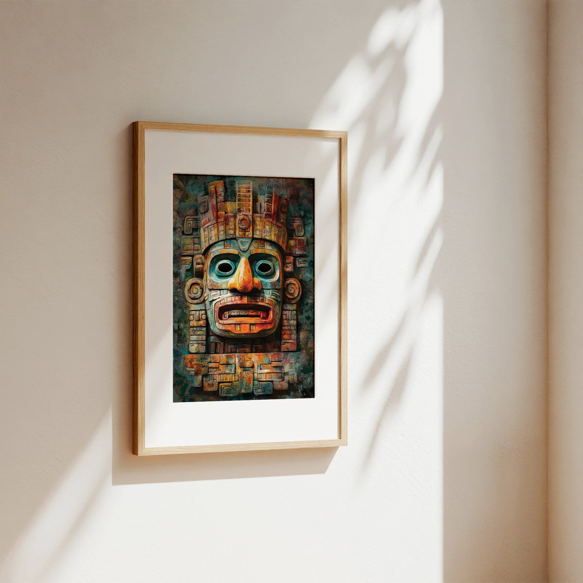 A framed art print featuring a colorful Aztec-style calendar mask, displayed on a warm beige wall with soft, dappled sunlight casting leaf shadows.