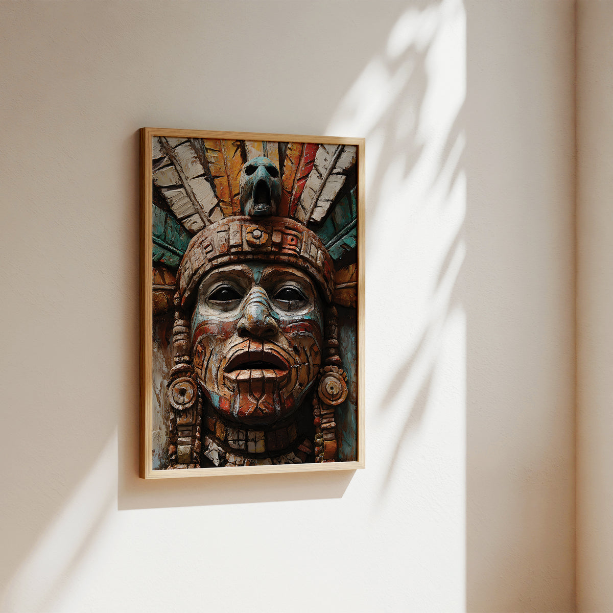 A framed art print featuring a detailed Aztec-style face sculpture with colorful painted details, hanging on a warm beige wall with soft sunlight and leaf shadows.