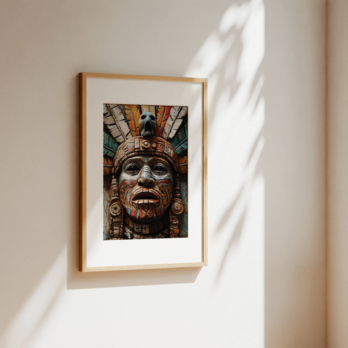 A framed art print featuring a detailed Aztec-style sculpture face, displayed on a warm beige wall with soft, dappled sunlight casting leaf shadows.