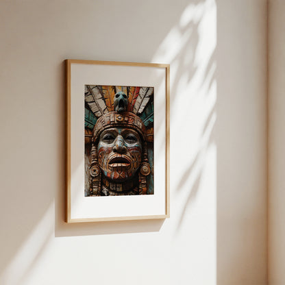 A framed art print featuring a detailed Aztec-style sculpture face, displayed on a warm beige wall with soft, dappled sunlight casting leaf shadows.