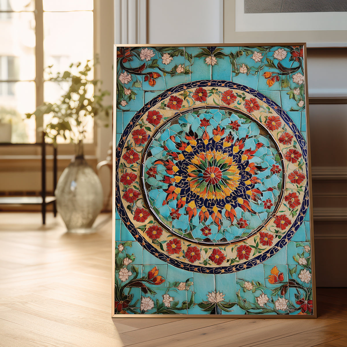 A framed decorative tile artwork leaning against a wall on a light wood floor in a bright interior. The artwork features a turquoise background with a large circular floral mandala and surrounding floral motifs in red, orange, yellow, green, and cream.