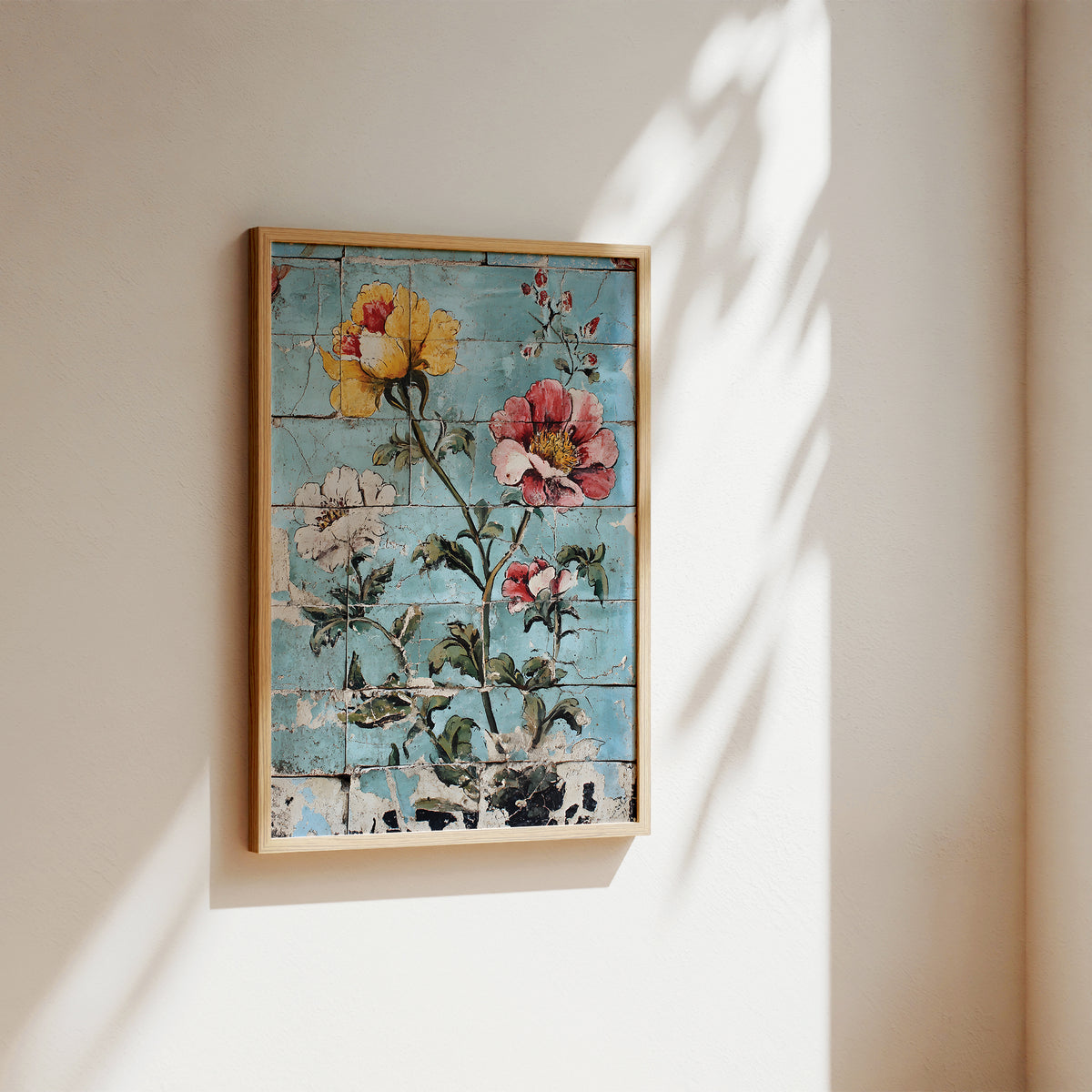 Framed wall art hanging on a beige wall, featuring a rustic floral design with yellow, pink, and white flowers on a distressed light blue background. The artwork is enclosed in a light wood frame and photographed in soft natural light with leafy shadows cast across the wall.